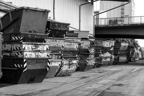 A row of large, industrial metal skips lined up along a concrete surface outside a warehouse or industrial building, with visible signs of rust and wear on the containers' surfaces. The skips are rectangular, open-topped, with some showing peeling paint and corrosion, suggesting frequent use for waste collection or disposal. The background includes a corrugated metal wall of the building and a raised platform or walkway with a guardrail, indicating an operational industrial setting. The area appears clean but utilitarian, suited for rubbish removal services in Lewisham or surrounding areas. The lighting is natural, casting shadows on the ground, emphasizing the textured surfaces of the metal skips and the concrete pavement. Slight vegetation and trees are faintly visible in the distant background, adding a subtle environmental context related to waste management outside urban or commercial premises. This scene depicts a typical setup for commercial waste collection with various containers ready for loading or collection by waste management specialists like Commercial Waste Lewisham.