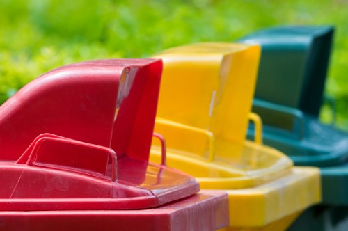 A row of three large, brightly coloured plastic wheelie bins positioned outdoors on a paved surface. The bin in the foreground is red, with a textured surface and a slightly raised lid, and has a sturdy grab handle on the front. To its right, a yellow bin features a smooth, glossy finish with a similar design and handle, while further to the right, a green bin is partially visible, with a matte finish and positioned in line behind the yellow bin. The background shows a grassy, green area, suggesting a residential or commercial outdoor space, typical of waste collection sites or waste management areas in Lewisham. The bins are used for general rubbish collection and are associated with waste removal services such as those provided by Commercial Waste Lewisham.