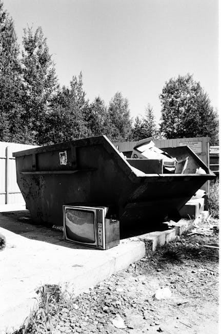 A large, black metal dumpster with a rectangular shape and high sides, positioned on a concrete paving slab in an outdoor area. The dumpster has a rough, weathered texture with visible rust and dirt, 